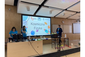 trzy osoby na scenie, w tle tablica z napisem 