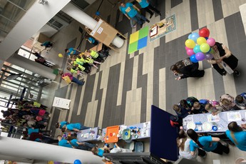 ludzie wchodzący na budynku, widok z góry na hol, w którym stoją przygotowane stoły z eksperymentami, przy jednym ze stołów dzieci przyglądają się pokazowi