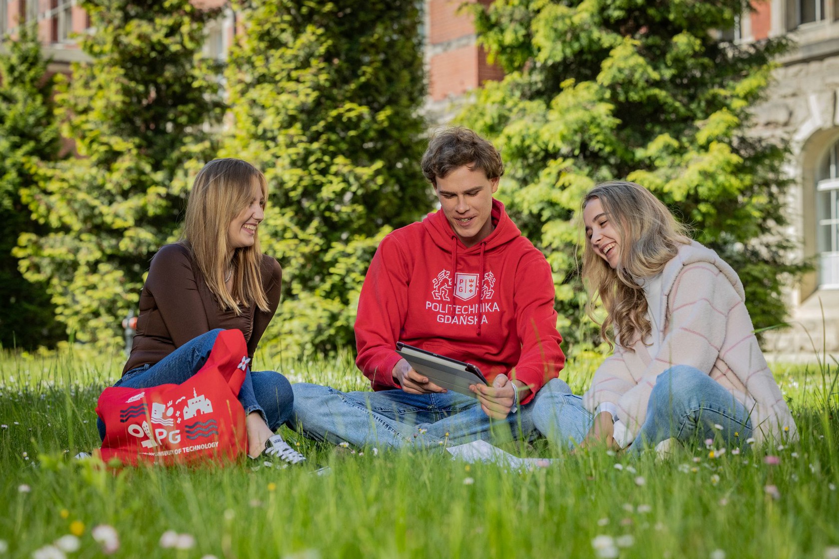 troje studentów siedzi na trawie przed Gmachem Głównym